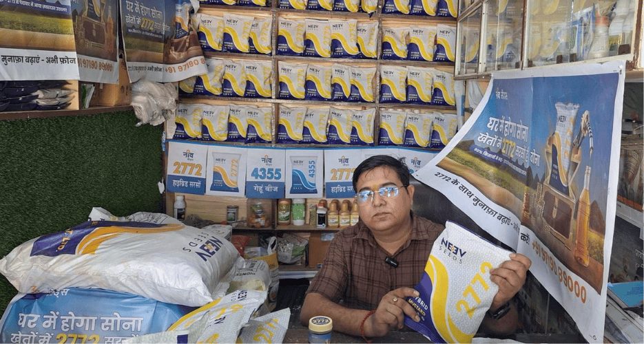 Seed retailer in his shop holding a Neev Seeds packet, with campaign posters