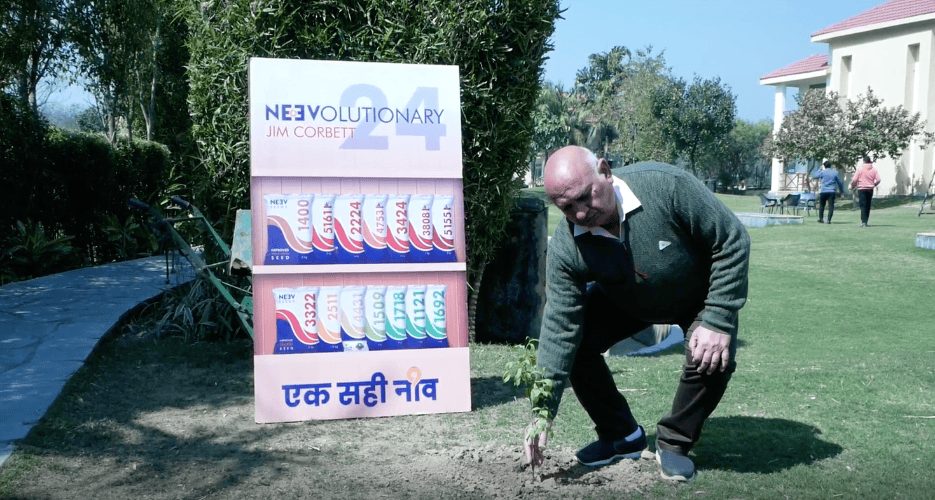 A guest planting a sapling beside a Neev Seeds standee on open grounds