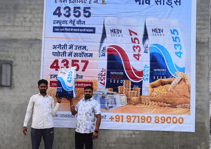 Two men in front of a large Neev Seeds wheat seed hoarding in a village